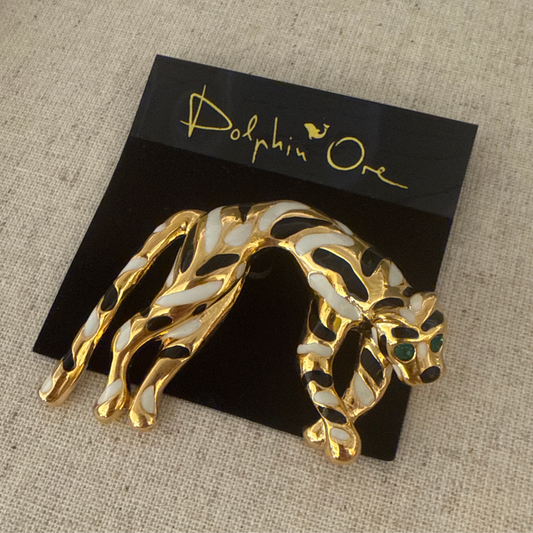 Gold and black animal-shaped brooch on a black card with 'Dolphin One' branding, placed on a textured beige surface.